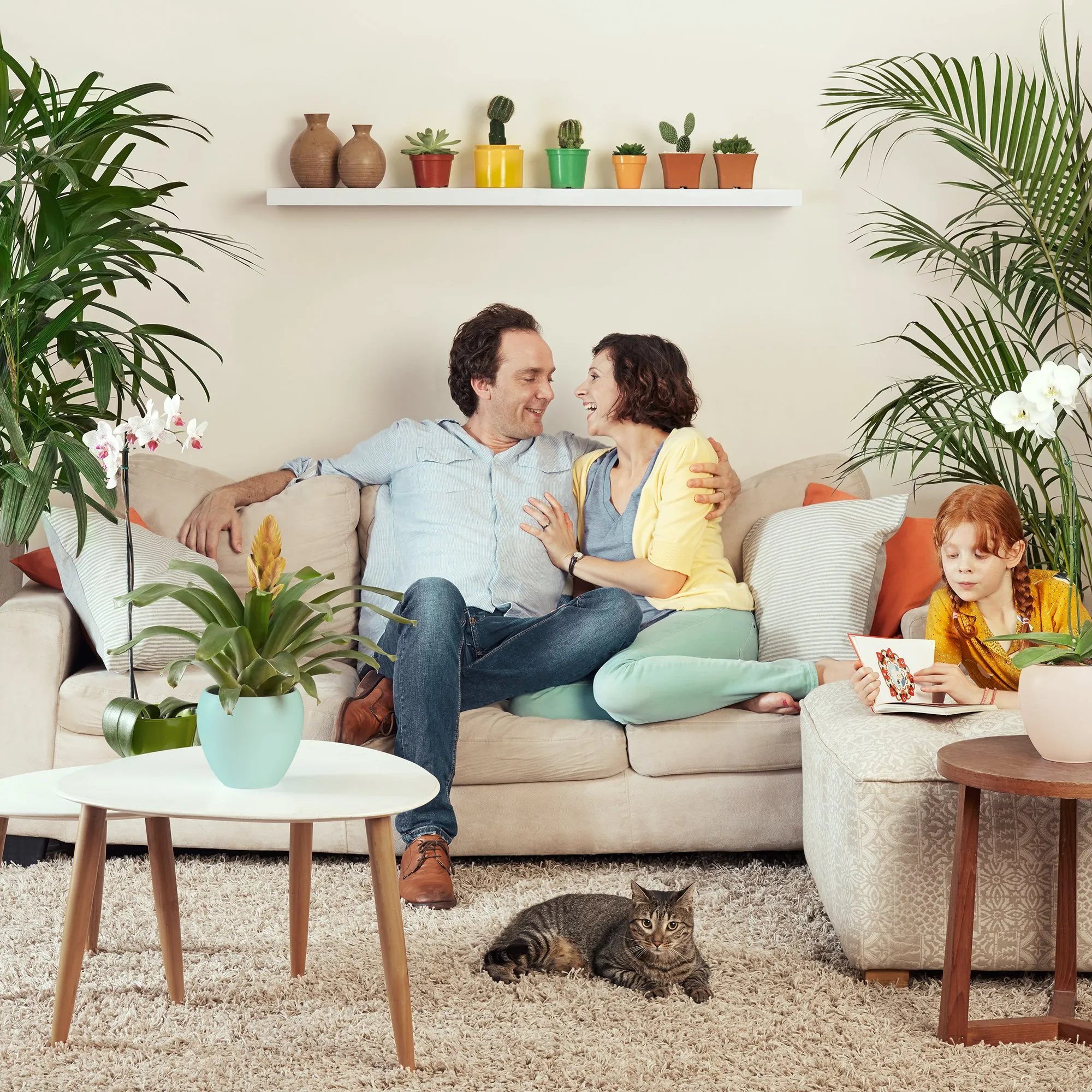 A happy family in the living room surrounded by plants and decorations, with a cat lying on the rug and a girl reading a book.