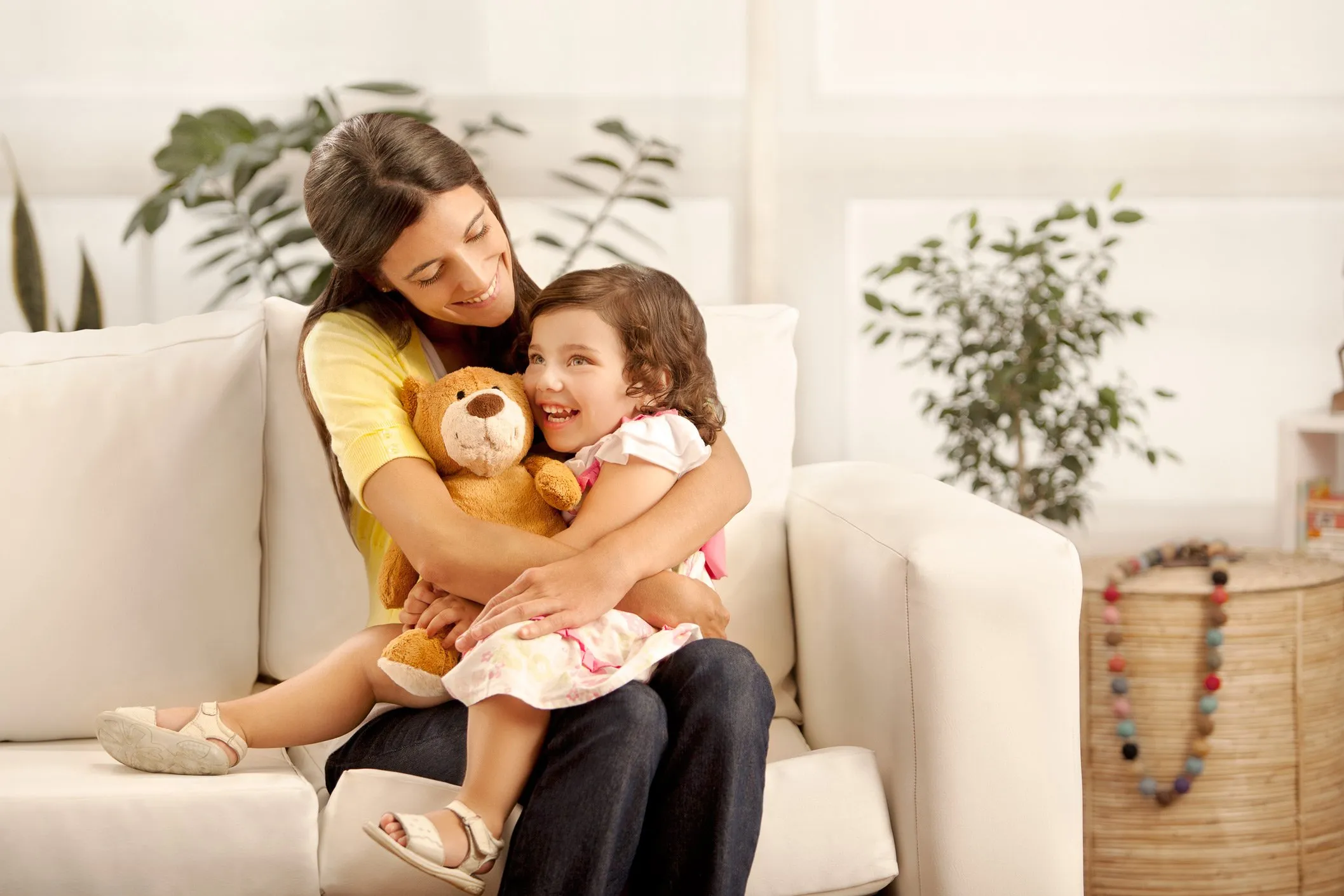 A mother and her young daughter hugging and smiling on a sofa, with the girl holding a teddy bear, in a homey setting with plants and decorations