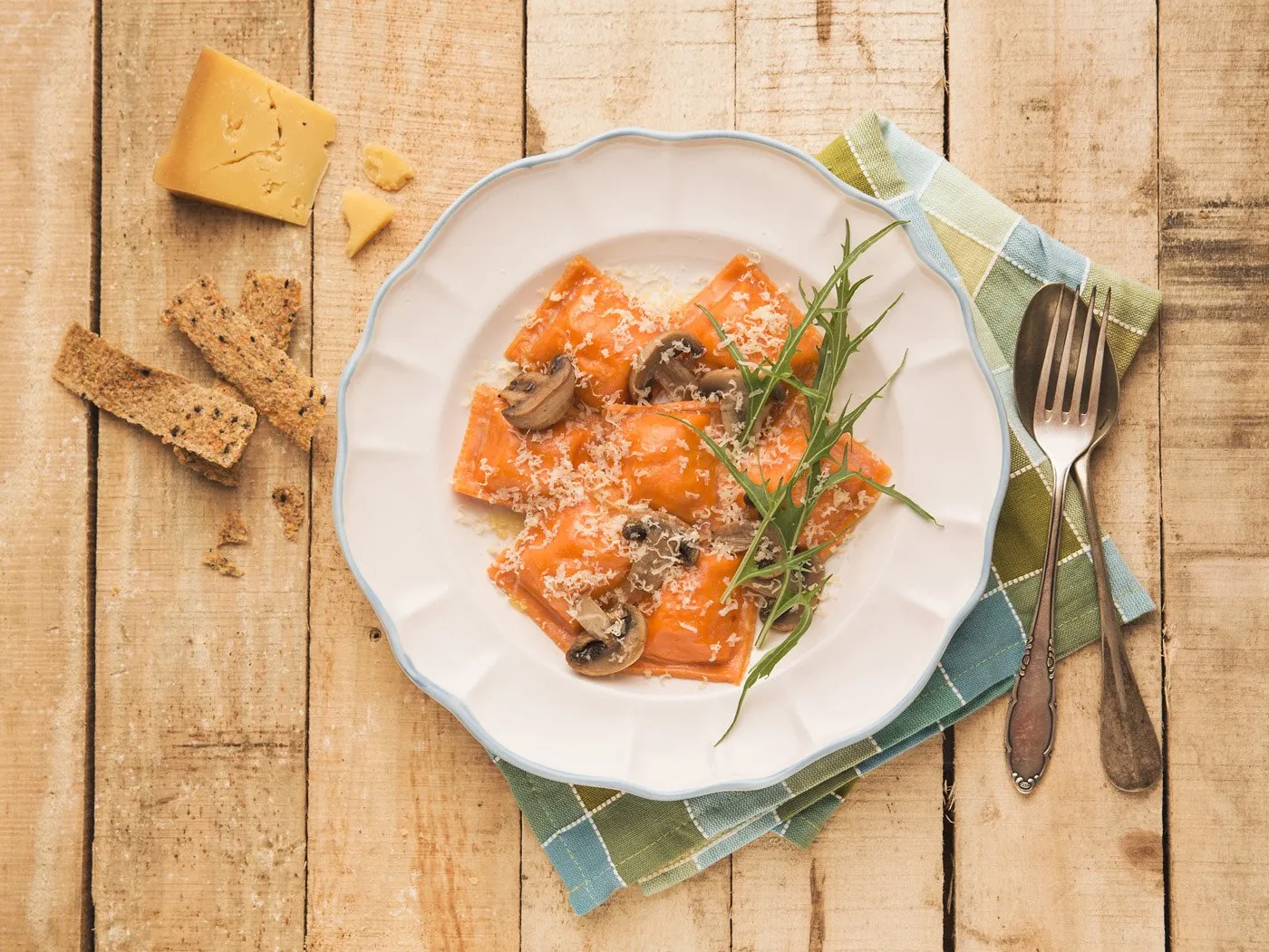 A pasta dish with tomato sauce, grated cheese, mushrooms, and arugula on a white plate on a wooden table, with a fork and spoon on a cloth. To the left, cheese, whole-grain bread, and crackers. An Italian meal.