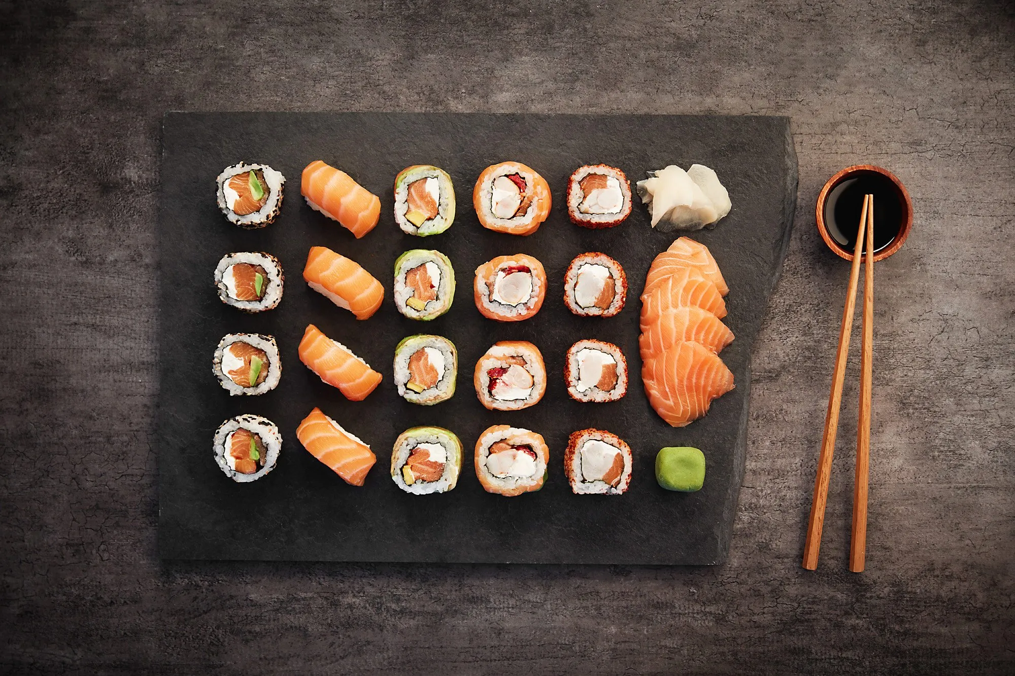 A plate with an assortment of sushi on a black tray, served with ginger, wasabi, and soy sauce in a bowl, with chopsticks.