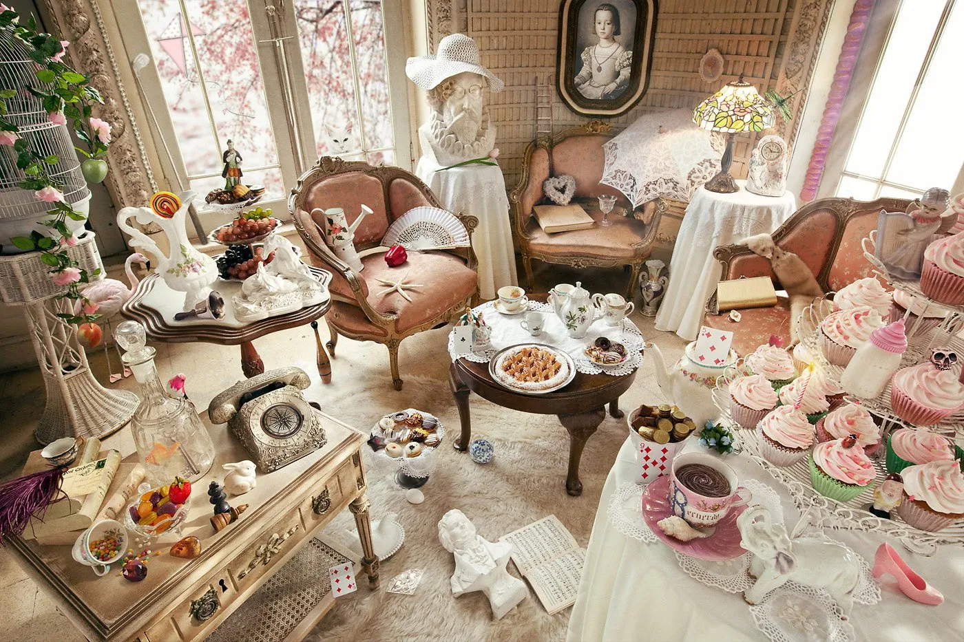 A room decorated with antique furniture and decorative objects, including teacups, pastries, pink cupcakes, porcelain, a typewriter, a ceramic dog figurine, and vintage decorations in pastel and pink tones.